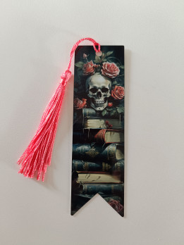 Marque-page illustré d'un crâne entouré de roses sur une pile de livres avec un pompon rose
