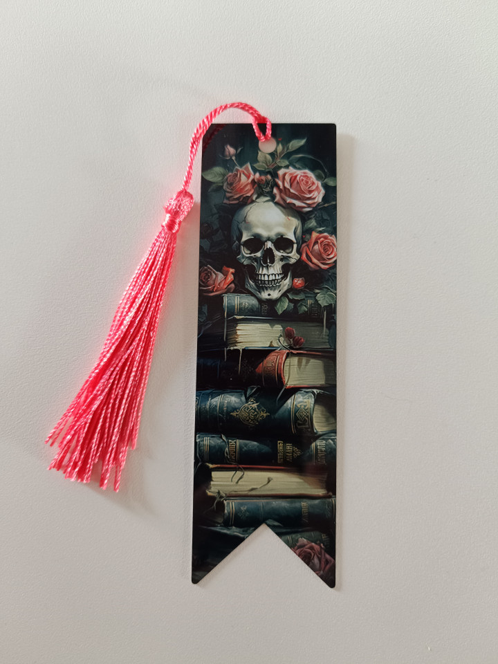Marque-page illustré d'un crâne entouré de roses sur une pile de livres avec un pompon rose