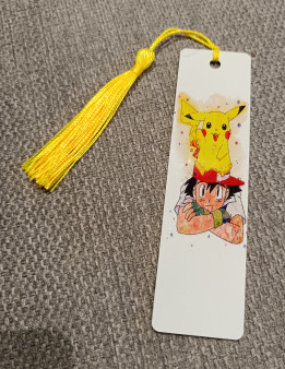 Marque-page blanc illustré d'un garçon à casquette rouge portant une créature jaune sur la tête, avec pompon jaune