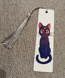 Marque-page blanc illustré d'un chat violet foncé avec un croissant de lune sur le front et pompon argenté