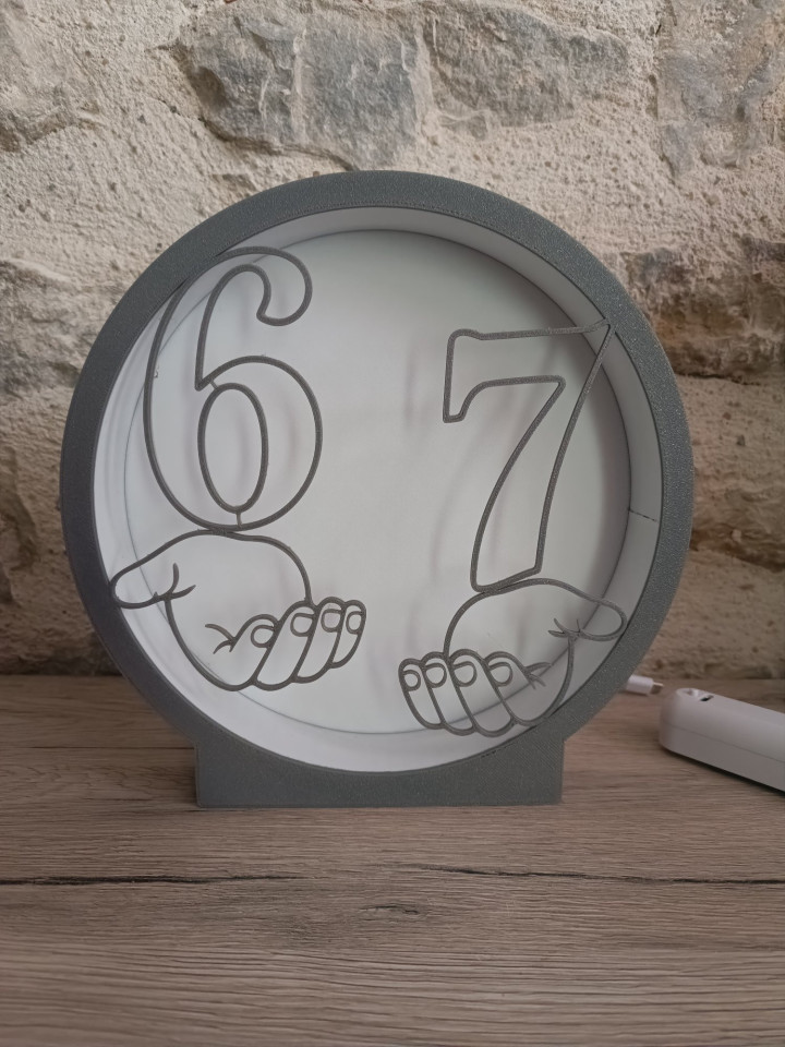 Veilleuse ronde noire illuminée montrant les chiffres 6 et 7 sur des mains stylisées avec contour LED rose et bleu